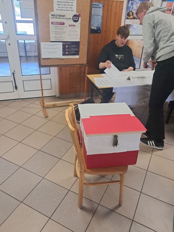 Uczeń pobiera kartę wyborczą od członka komisji, a na pierwszym planie widać urnę wyborczą