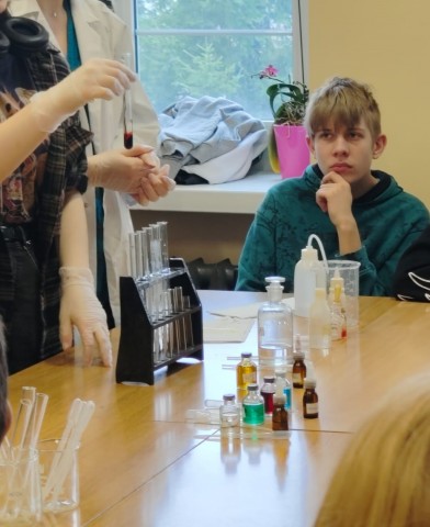 Kadr z warsztatów chemicznych. Zdjęcie przedstawia odczynniki, substancje i obserwującego doświadczenie ucznia. 
