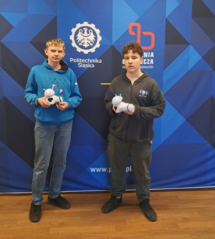 Zdjęcie przedstawia 2 laureatów XV edycji Międzynarodowego Konkursu Robotic Tournament, stoją na ściance reklamującej konkurs i trzymają w rękach roboty Photon